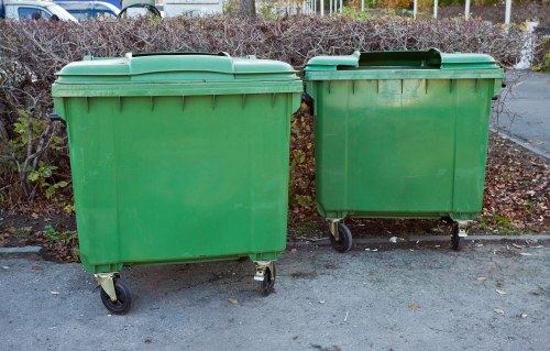 Two large green wheelie bins made of durable plastic with smooth surfaces and solid lids, positioned side by side on a paved area. The bin on the left is slightly larger with four small wheels visible at the base, while the bin on the right is smaller, also with four wheels, and appears to be used for general rubbish collection. Both bins are placed facing forward, with the waste containment area at the top, and are situated on a concrete or asphalt surface, slightly weathered and with some ground debris visible nearby. In the background, there is a row of dense bushes without foliage, indicating an outdoor environment possibly adjacent to residential grounds. The scene is well-lit, capturing the neutral tones of the bins and their surroundings, aligning with waste removal services offered by House Clearance Marlow in the local area.