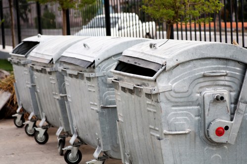 A row of four large, industrial-style metal wheelie bins with hinged lids, positioned on a paved pavement in an outdoor urban environment. Each bin has a textured, galvanized steel finish, with visible ridges and a slightly weathered appearance. The lids are slightly open, revealing dark interiors, and are equipped with metal handles on the front. The bins are aligned in a straight line, with small wheels at the base for mobility, and are situated near a metal fence and parked vehicles in the background, suggesting a commercial or residential area in Marlow. The scene is evenly lit, capturing the utilitarian nature of rubbish collection containers used by local waste management services like House Clearance Marlow. This detailed visual description supports understanding of typical refuse storage used in household or property clearance projects within the town, emphasizing the importance of proper waste disposal equipment in effective rubbish removal services.