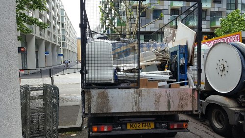 A flatbed truck parked on a city street loaded with a variety of household and construction rubbish, including white plastic furniture, a blue and white appliance, cardboard boxes, and miscellaneous debris, with a metal fencing panel on top. The truck's rear is rusted and slightly weathered, and the vehicle is positioned on a pavement adjacent to a modern residential or commercial building with balconies and large windows. In the background, urban elements such as traffic lights, a pedestrian crossing, and other city infrastructure are visible. This scene illustrates the type of rubbish clearance services provided by House Clearance Marlow in the local area near Marlow, highlighting a typical load of household or small-scale building waste ready for removal and disposal.