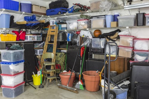 A cluttered storage area or garage showing multiple plastic storage boxes stacked and lined along the metal shelving unit, with some boxes featuring visible red and grey lids, and others transparent revealing contents inside. Various household items such as a rolled-up rug, a wooden ladder, and gardening tools including brooms and a mop are leaning against the shelving, with some stacked chairs and a small folding step stool nearby. Several empty flowerpots and a hand truck or trolley with a metal frame are positioned among the objects, while the overall scene exhibits a busy, disorganized space suitable for rubbish removal or clearance services in the Marlow area. The environment is brightly lit, highlighting the mixture of materials like plastic, wood, and metal, typical of a residential garage or storage room used for household and garden waste accumulation. House Clearance Marlow specializes in clearing such clutter efficiently, helping to tidy these types of spaces in the local postcode district.
