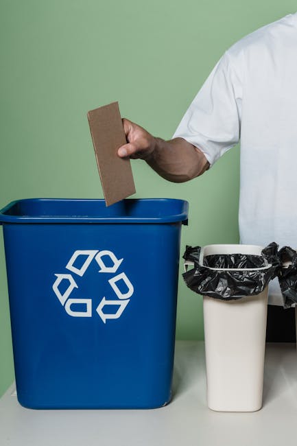 A person is seen disposing of crumpled brown paper bags into a grey rubbish bin with a white lid, situated on a wooden surface indoors. The person's arm extends over the bin, holding a paper bag ready
