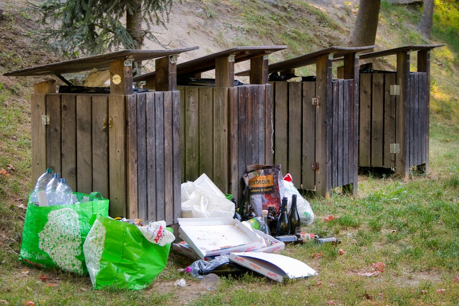The image shows four wooden outdoor rubbish bins with sloped roofs situated on a grassy area next to a hillside with trees in the background. The bins are weathered, with dark brown stains and visible